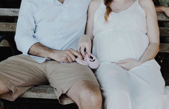 Close up of man and woman sitting on a bench holding small knitted shoes, and the woman holds her baby bump