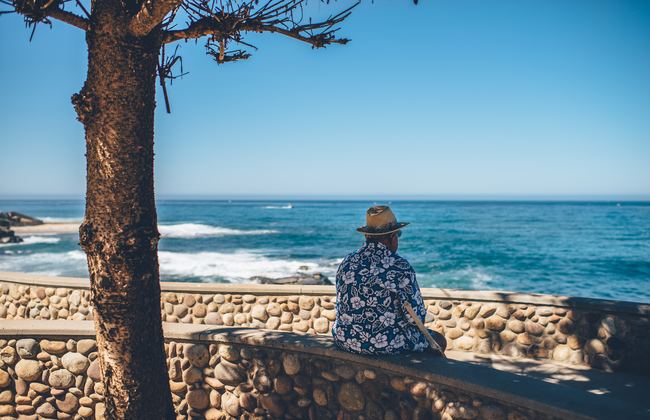 Man wearing a hat sitting on a wall by the beach, looking out to sea